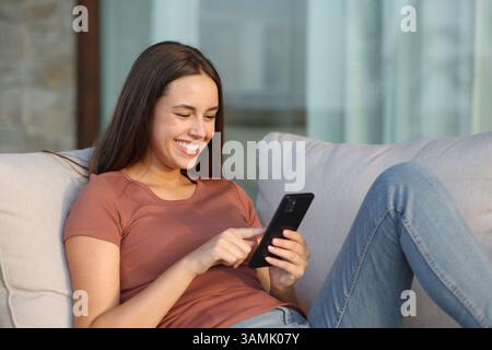 Happy woman sitting in a terrace sofa is using smart phone at home or hotel Stock Photo