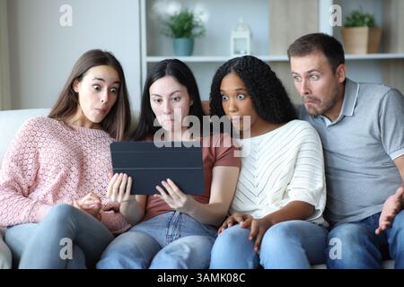 Perplexed group of friends checking tablet sitting on a couch at home Stock Photo
