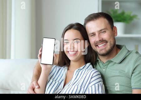 Happy couple at home showing blank mobile phone screen sitting on a couch looking at camera Stock Photo