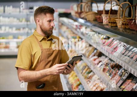 Bearded store employee wearing apron checking inventory on digital device while standing in front of products in grocery store aisle during work shift Stock Photo