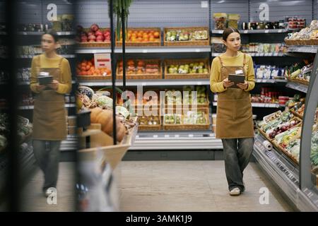 Grocery store employee wearing apron and holding clipboard while checking inventory in produce section. Various fruits and vegetables displayed in background Stock Photo