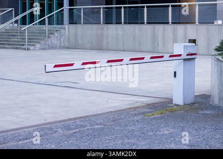 Close-up view of a white and red striped security barrier gate lowered across a paved entrance near a modern building, symbolizing controlled access a Stock Photo