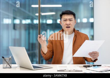 An asian businessman looks stressed holding papers, possibly reviewing documents, with a perplexed expression in a modern office. Stock Photo