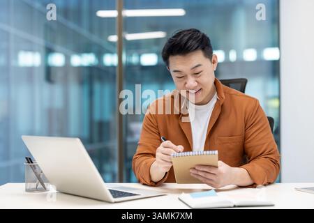 An asian businessman sits at his desk, making notes in a notebook while working on a laptop in a modern office setting. Stock Photo