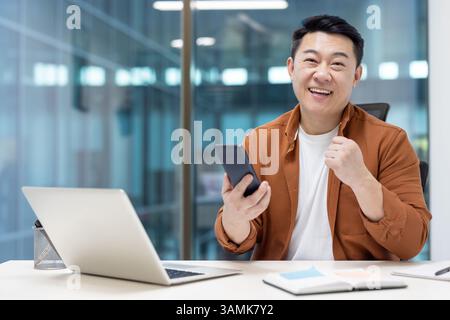 An Asian businessman celebrates a success in his office, holding a phone with excitement. The man is dressed casually. Stock Photo