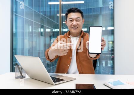 An asian businessman smiles and points at his smartphone with blank screen, showing its features. Stock Photo