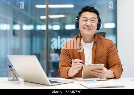 An asian businessman smiles widely while taking notes, wearing headphones, and using his laptop at work. He looks content. Stock Photo