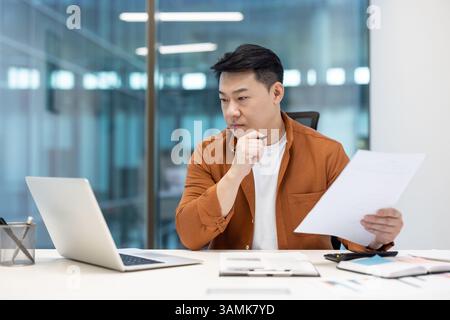 Asian businessman in office, looking thoughtfully at documents, considering data and making decisions. Analyzing business reports at his desk. Stock Photo