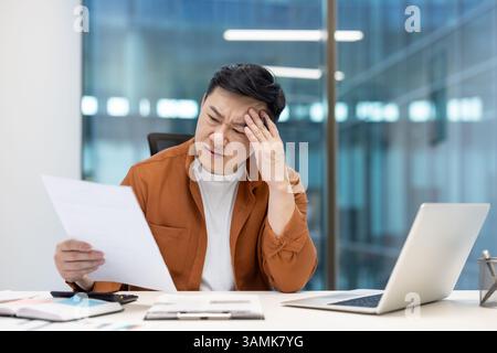 An asian businessman is working at his desk, appearing stressed while reviewing financial documents and data with a concerned look. Stock Photo