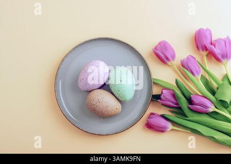 Easter composition with speckled pastel eggs on plate and pink tulips on beige background. Top view, flat lay Stock Photo