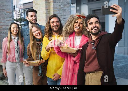 Multiracial group of happy young adults enjoying street noodles and taking a selfie together in an urban outdoor environment Stock Photo