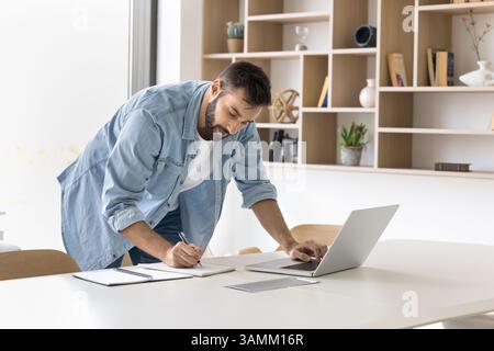 Focused millennial man stand by workplace browse internet write notes Stock Photo