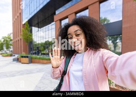 A young, smiling woman with curly hair takes a selfie, waving hello against an urban backdrop of modern architecture. Stock Photo