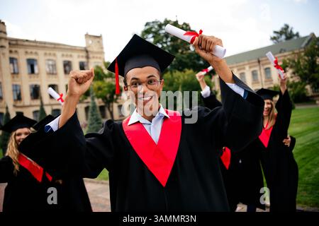 Joyful young graduates celebrating their achievement in academic robes on a sunny day outdoors at a university campus. Stock Photo