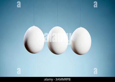 Three white eggs suspended in a line against a blue gradient background that ranges from light to dark. A minimalist composition. Stock Photo