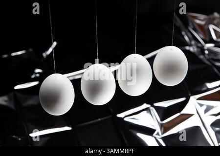 Four white eggs suspended on thin wires in front of a crumpled, reflective metallic background. Stock Photo
