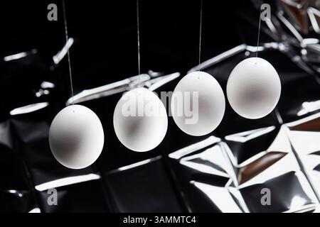 Four white eggs hanging in a slightly staggered horizontal line from thin, visible threads against a dark, crumpled, reflective foil background. Stock Photo