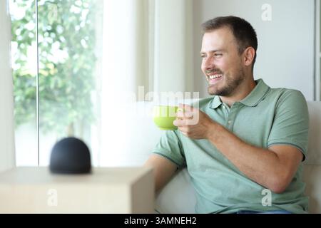 Happy man drinking coffee relaxing with smart speaker sitting on a couch at home Stock Photo