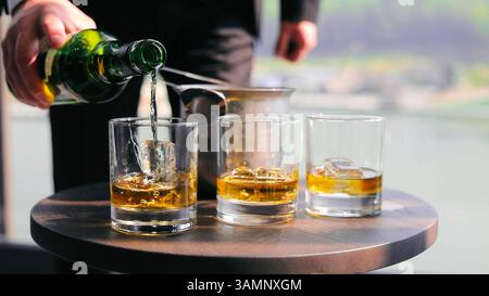 Bartender pouring whiskey into glasses with ice. A bartender's hand is seen pouring whiskey from a green bottle into glasses filled with ice cubes on Stock Photo