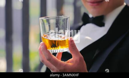 Elegant Man Holding a Glass of Whiskey in Formal Attire. A close-up of a man in formal attire, wearing a tuxedo and holding a glass of whiskey with Stock Photo