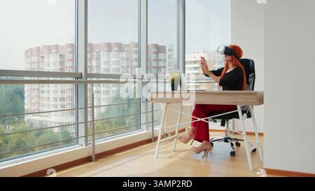Woman Using VR Headset in Modern Office. A woman wearing a virtual reality headset while seated at a desk in a bright, modern office overlooking city Stock Photo