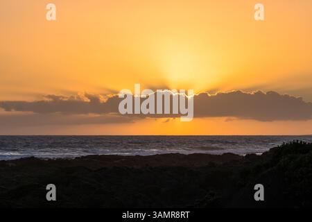 sunset, sun and rays behind a single long linear cloud in an orange sky showing the horizon and the sea or ocean, in the foreground the dark shoreline Stock Photo