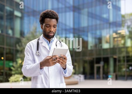 An african-american doctor reviews information on a tablet while outside a modern medical facility building, wearing his lab coat. Stock Photo