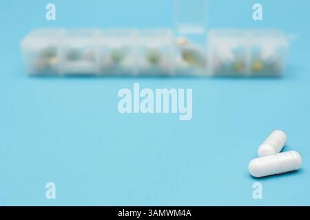 Two white capsules lie on a blue surface with a weekly pill case blurred in the background. pharmacy, daily medication, copy space Stock Photo