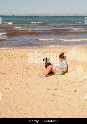 A woman sits reading on the beach at Les Meravelles whilst her pet labrador looks out on the Bay of Palma, Island of Mallorca, Spain Stock Photo