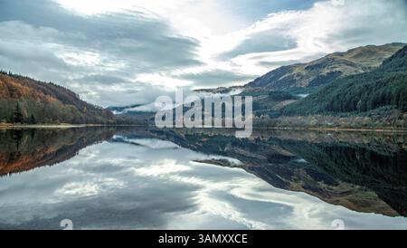 Low mist clinging to the mountainside and reflections of autumnal forests on the bank, in the tranquil waters of Loch Goilhead. Stock Photo