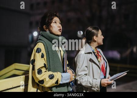 Two women having a conversation outdoors at night, one dressed in a scarf and the other holding a notebook Stock Photo
