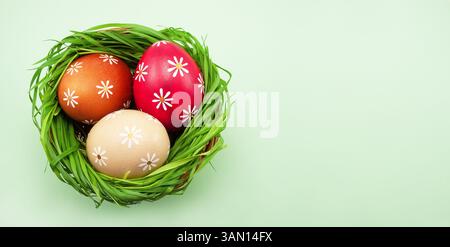 Colored Easter eggs in a basket with green grass on a light green colored table, top view. Stock Photo