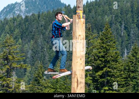 A lumberjack, dressed in a blue plaid shirt and red suspenders, competes in a lumberjack competition. Standing on a narrow platform using an axe. Stock Photo