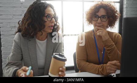 Hispanic women discussing work in an office interior, showcasing a diverse professional environment with coffee culture and teamwork dynamics. Stock Photo