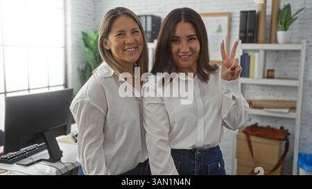 Two smiling hispanic women stand in a bright office, one making a peace sign, embodying teamwork and confidence in a professional setting. Stock Photo