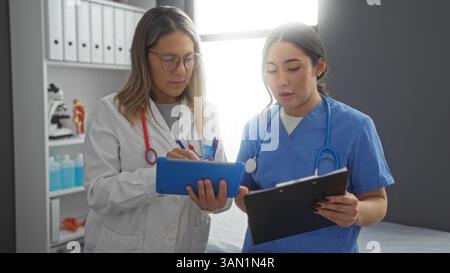 Women doctors collaborating in hospital room discussing patient care with electronic tablet and clipboard in professional medical setting. Stock Photo