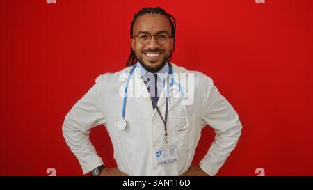 African american male doctor with braids and glasses smiling confidently against a vivid red background in medical attire holding a stethoscope. Stock Photo
