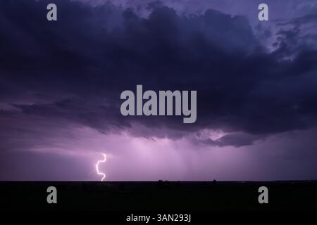A powerful lightning bolt cuts through the night sky during a thunderstorm, casting a violet glow over the landscape. Stock Photo