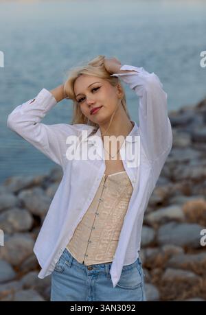 a portrait of a young woman posing outdoors near a body of water with a rocky shoreline. She has blonde hair and is wearing a stylish outfit consistin Stock Photo