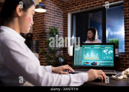 Female business analyst using her personal computer to examine marketing trends and track company performance in brick wall office. Asian employee holding her mouse as she scrolls through her laptop. Stock Photo