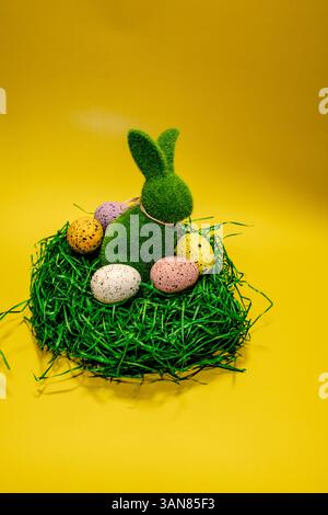 Decorative green bunny surrounded by colorful Easter eggs in a nest on a vibrant yellow background Stock Photo