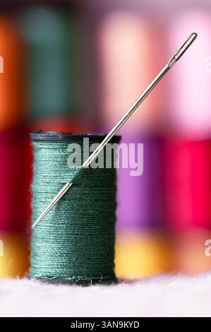A spool of green thread with a sewing needle through it, colorful spools stacked up in the background Stock Photo