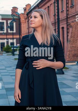 Young pregnant woman in a black dress poses confidently outside historic red brick buildings in a town square during sunset Stock Photo
