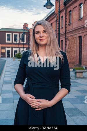 Young pregnant woman in a black dress poses confidently outside historic red brick buildings in a town square during sunset Stock Photo