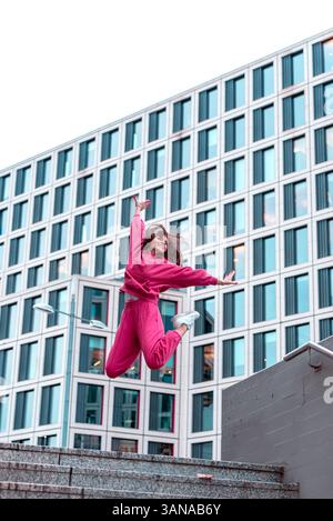 Young woman in pink sports outfit smiling and jumping in front of an office building. Stock Photo