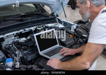 Engineer bearded man looking at inspection vehicle details under car hood using laptop Stock Photo