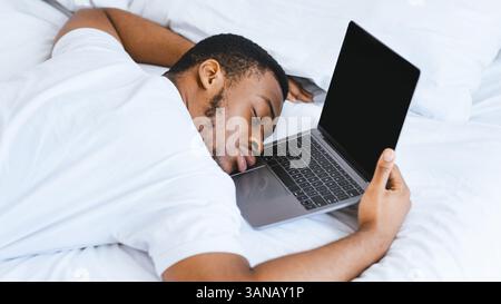 Exhausted Black Guy Sleeping On Laptop Lying In Bed At Home Stock Photo
