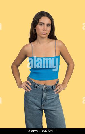 Confident young woman standing with hands on her hips, exuding strength and style against a vibrant yellow backdrop Stock Photo