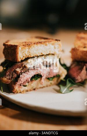 A sandwich with meat and greens is cut in half and placed on a white plate. The sandwich is served on a wooden table Stock Photo