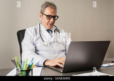 Senior male doctor using laptop during online consultation or medical work in a healthcare office, concept of digital healthcare and telemedicine. Stock Photo